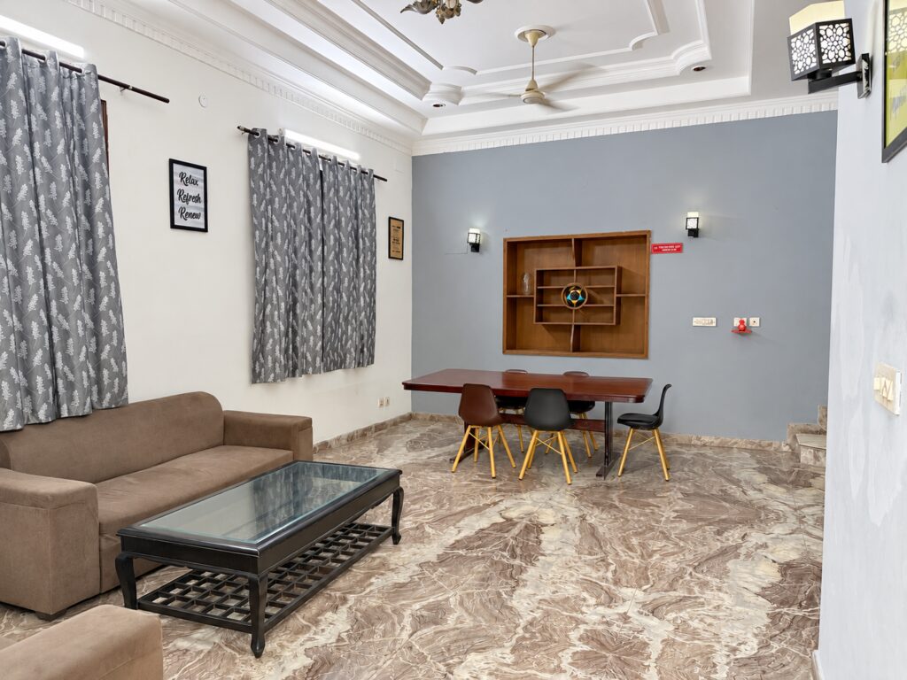 Shared lounge with brown sofa, patterned curtains and dining table at GableStay girls’ PG